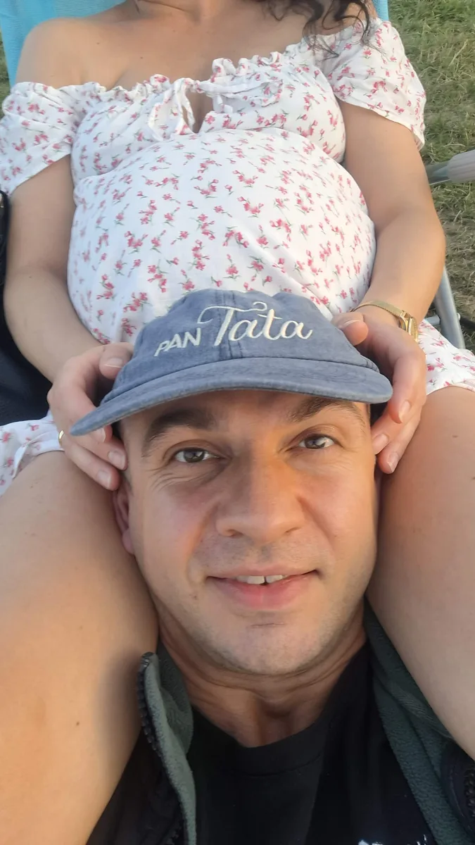 Michał Czekaj leży z głową na kolanach Anny w trzecim trymestrze ciąży, a ona wkłada mu na głowę dżinsową czapkę z haftem „Pan Tata” — wakacyjne popołudnie nad zalewem